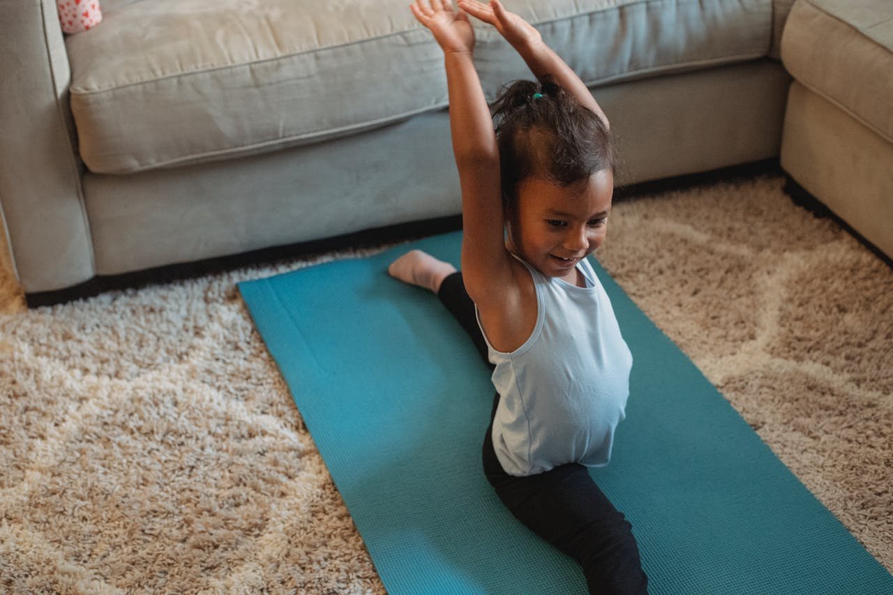 From above of cheerful little ethnic girl in sportswear doing leg split on stretch mat with raised hands at home